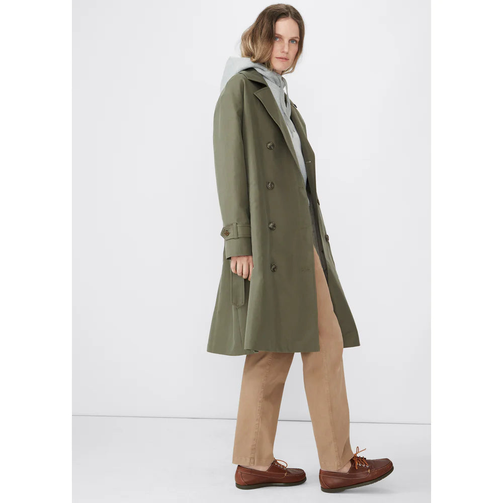 A person with long hair is wearing the chic Rox trench by Kule, made from water-resistant olive green fabric, over a gray hoodie and beige pants. Standing against a plain white background, they have brown shoes on and are gazing to the side.