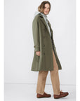 A person with long hair is wearing the chic Rox trench by Kule, made from water-resistant olive green fabric, over a gray hoodie and beige pants. Standing against a plain white background, they have brown shoes on and are gazing to the side.