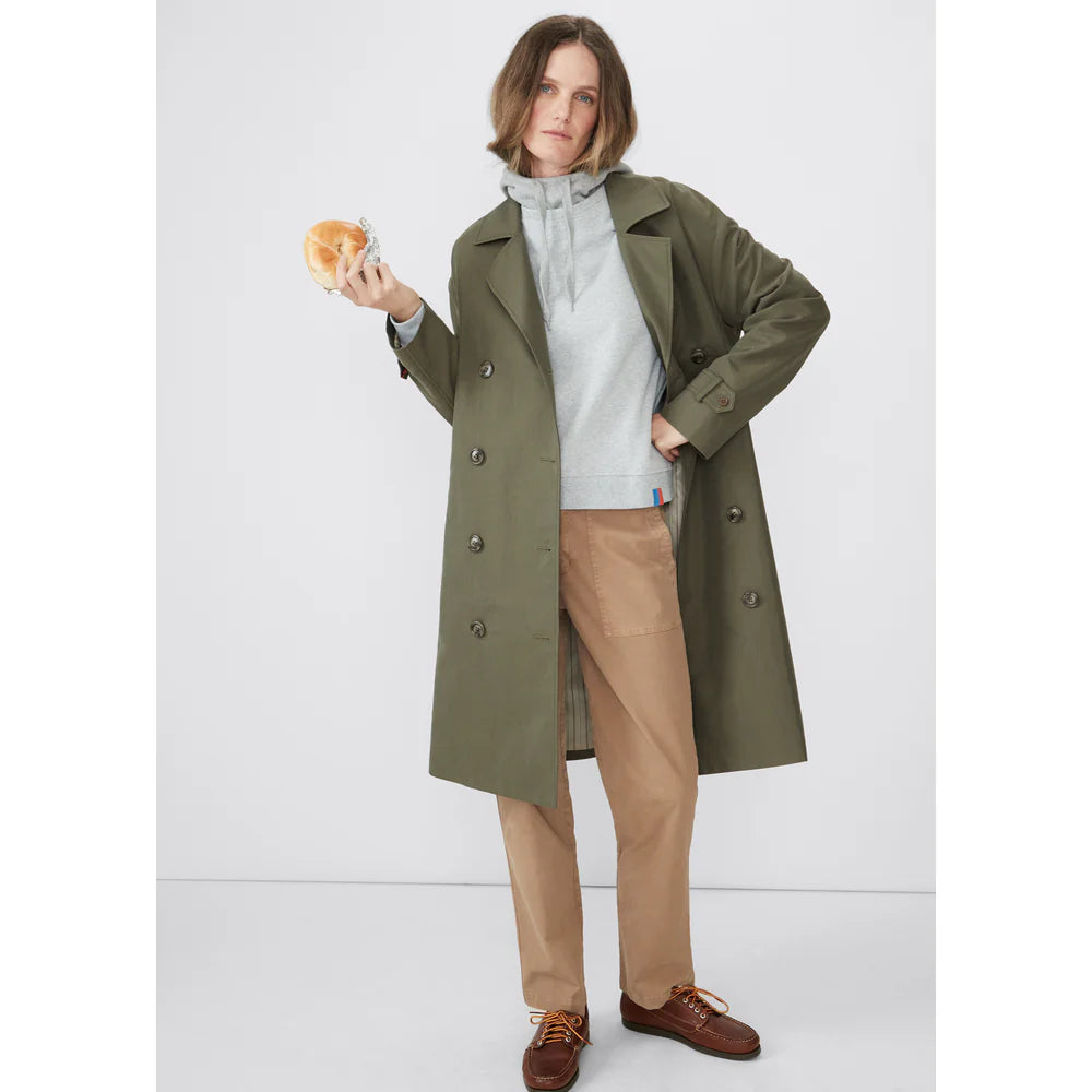 A person with medium-length hair stands against a plain background, wearing The Rox trench by Kule—a chic, olive green oversized trench coat—over a gray hoodie and beige pants, holding a bagel in one hand. Their expression is neutral.