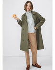 A person with medium-length hair stands against a plain background, wearing The Rox trench by Kule—a chic, olive green oversized trench coat—over a gray hoodie and beige pants, holding a bagel in one hand. Their expression is neutral.