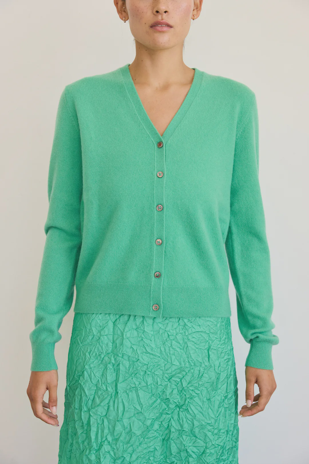Someone wears the Brazeau Tricot Minlan Cardigan in mojito-green cashmere with a V-neck and a textured green skirt against a plain light background, their face partially visible.