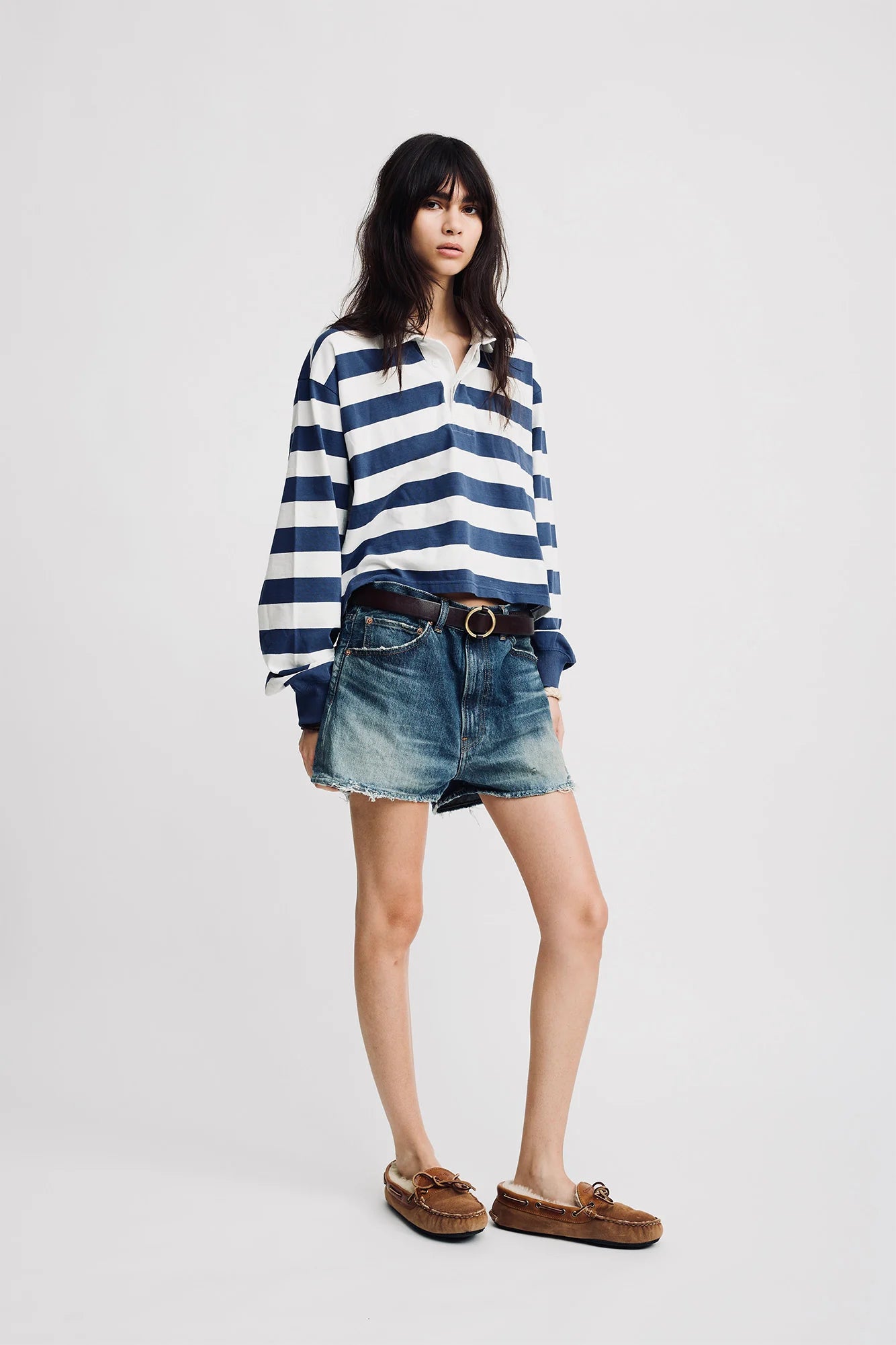 A person with long dark hair and a neutral expression wears the Denimist Rugby LS Tee in blue ecru stripe, paired with faded denim shorts, a brown belt, and brown loafers, standing against a plain background.