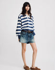 A person with long dark hair and a neutral expression wears the Denimist Rugby LS Tee in blue ecru stripe, paired with faded denim shorts, a brown belt, and brown loafers, standing against a plain background.