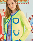 Someone with long hair wears a cotton crochet lime green Hunter Bell NYC Russell Cardigan with blue trim and pockets over a blue and pink striped dress. Neon yellow and blue geometric artwork decorates the wall behind.