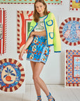 A woman is in a vibrant room wearing a Russell Cardigan by Hunter Bell NYC in neon yellow and green. Her outfit includes a printed crop top, a blue patterned skirt, and blue sandals. The backdrop showcases lively, patterned art pieces on the wall.