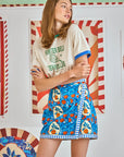 A woman with long hair wears a "Hunter Bell NYC Tennis Club" t-shirt and a Powell Skirt in vibrant orange grove print. The backdrop bursts with colorful, patterned artwork featuring bold stripes and geometric designs, enhancing the lively scene.