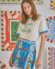 In front of vibrant, patterned backdrops, a young woman gazes at the camera with loose brown hair. She wears a white T-shirt with green text and Hunter Bell NYC's Powell Skirt in bright blue stretch sateen, featuring an Orange Grove print.