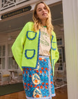 A woman in a neon green cardigan with blue trim, a graphic tee, and a Hunter Bell NYC Powell Skirt in vibrant Orange Grove print stands outside The Gasparilla Inn. She smiles with eyes closed, exuding relaxation.