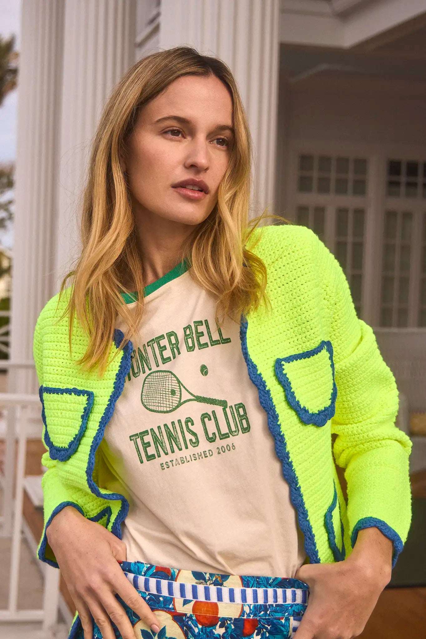 A woman with long blonde hair wears a neon yellow knit cardigan with blue trim over a Hunter Bell NYC Tennis Club Tee, standing in front of a white columned porch.