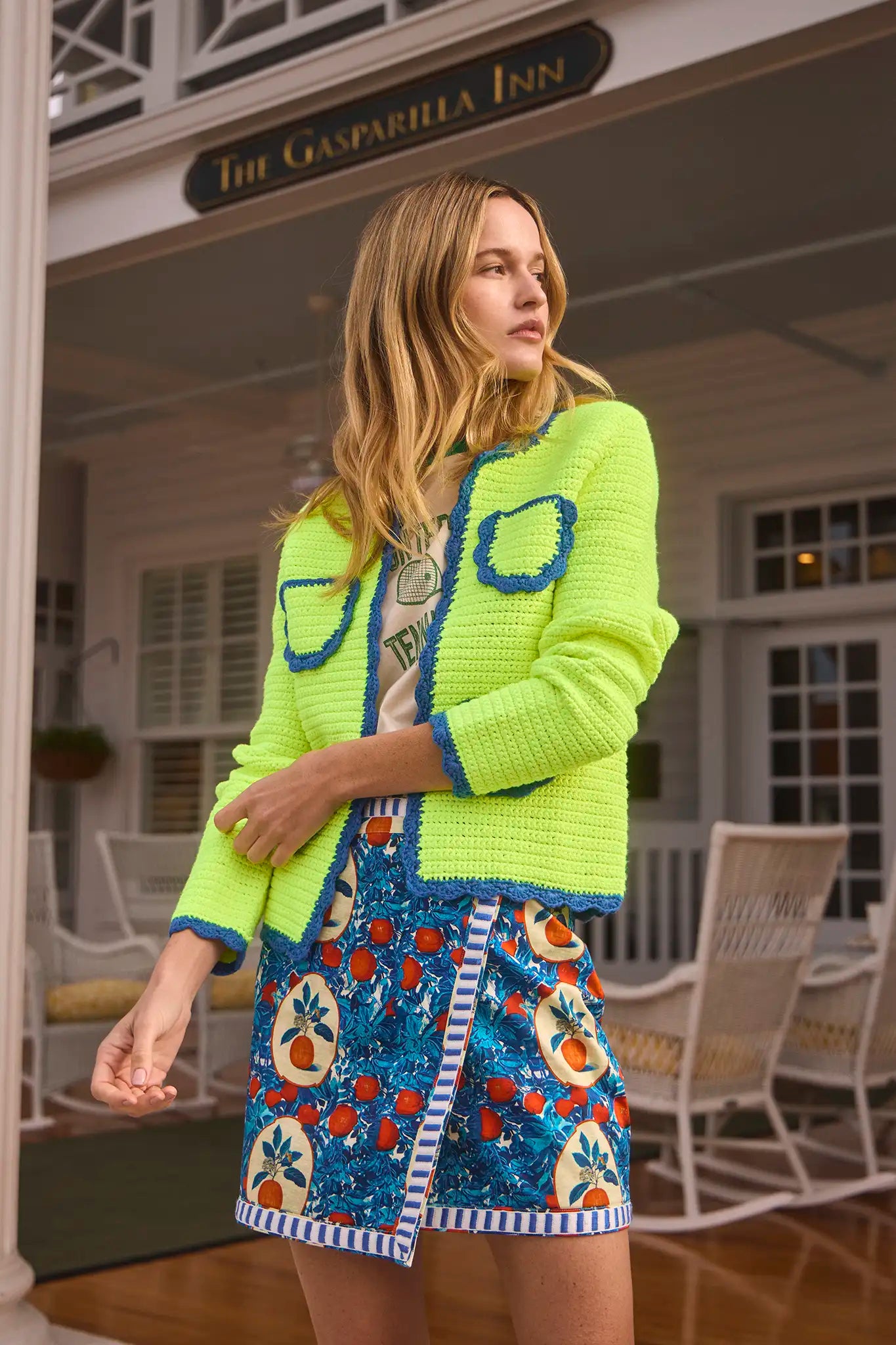 A woman confidently wears a Hunter Bell NYC Russell Cardigan over a T-shirt and a colorful fruit-patterned skirt. Her vibrant outfit stands out against the building with "The Gasparilla Inn" sign behind her.