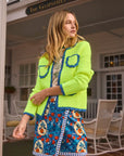 A woman confidently wears a Hunter Bell NYC Russell Cardigan over a T-shirt and a colorful fruit-patterned skirt. Her vibrant outfit stands out against the building with "The Gasparilla Inn" sign behind her.