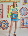 A woman in a vibrant space wears a Hunter Bell NYC Russell Cardigan with crochet trim, a white t-shirt, and a blue floral skirt. Neon yellow and blue sandals finish her look. The wall features an array of patterned artworks, creating a lively atmosphere.