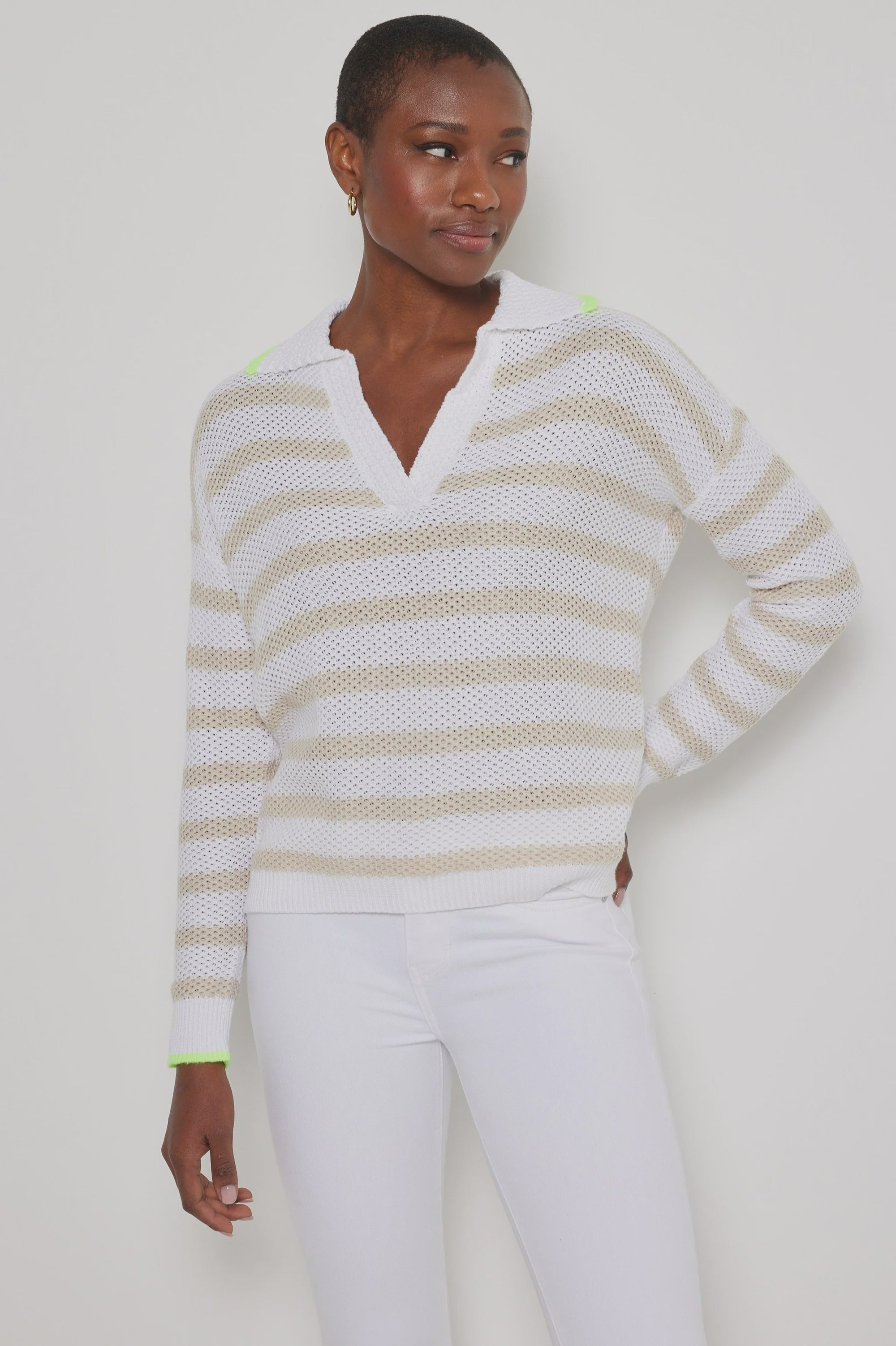 Against a light background, a person wears the Lisa Todd "Stripescape," a white and beige V-neck cotton sweater with neon green accents, teamed with white pants. The textured knit adds depth to their look, while their short hair and relaxed expression complete the image.