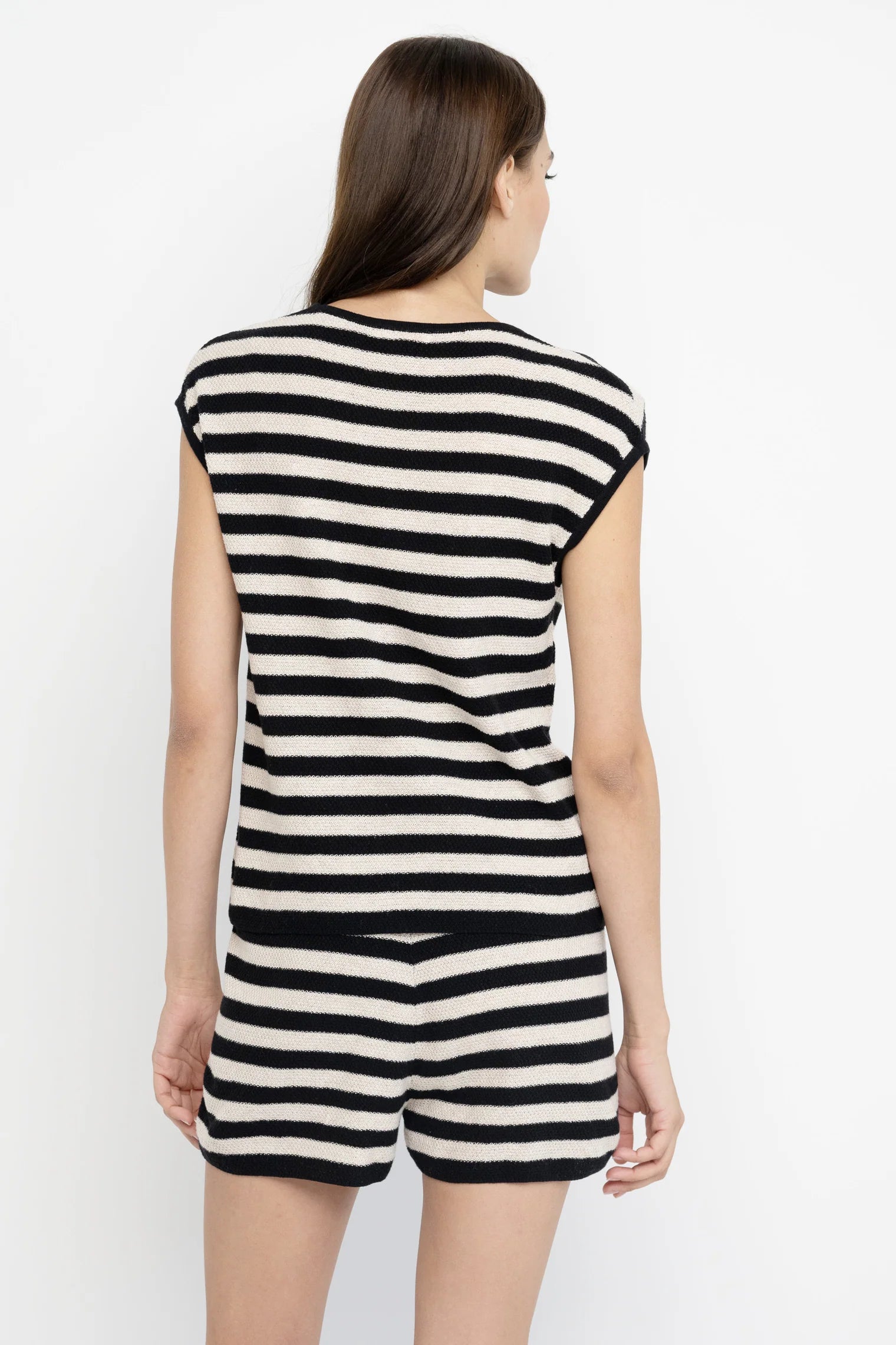 A woman with straight brown hair faces away, wearing the Lisa Todd Summer Lovin Sweater—a sleeveless crew neck black and white striped knit top with matching shorts. The background is plain white.