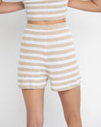 A model wearing the Lisa Todd Summer Lovin Short, featuring an elastic waist and tan and white horizontal stripes, paired with a matching crop top, stands against a plain white background with partially visible arms.