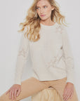 A person with long, wavy blonde hair sits on a wooden cube wearing the Lisa Todd "Star Dust," a light beige cashmere sweater with intarsia star patterns, paired with light brown pants. A plain white background accentuates the attire's understated luxury.