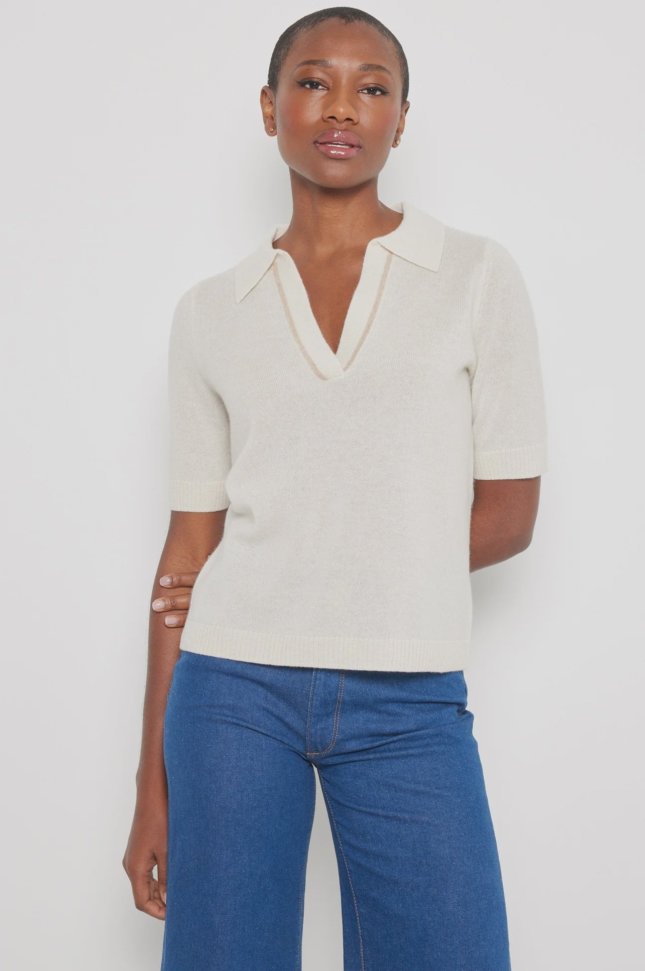 Against a white backdrop, an individual with short hair and a calm expression wears the Lisa Todd "Members Only" luxury knit polo sweater paired with blue jeans, one arm resting by their side.