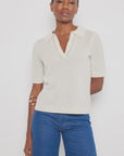 Against a white backdrop, an individual with short hair and a calm expression wears the Lisa Todd "Members Only" luxury knit polo sweater paired with blue jeans, one arm resting by their side.