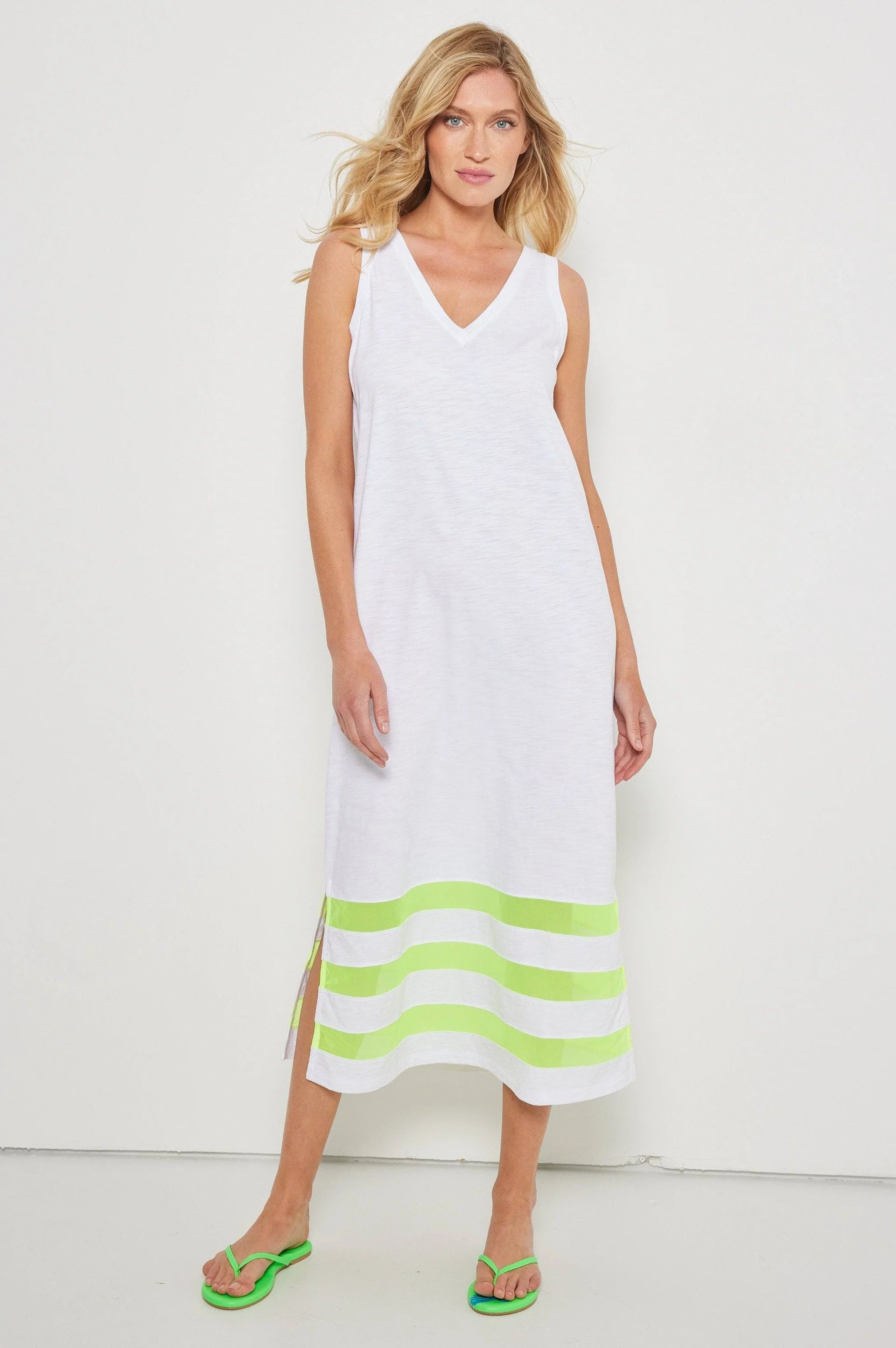 A woman models Lisa Todd's Best Dressed—a sleeveless white maxi dress with a v-neck and lime green stripes at the hem, paired with matching green flip-flops—against a plain white background.