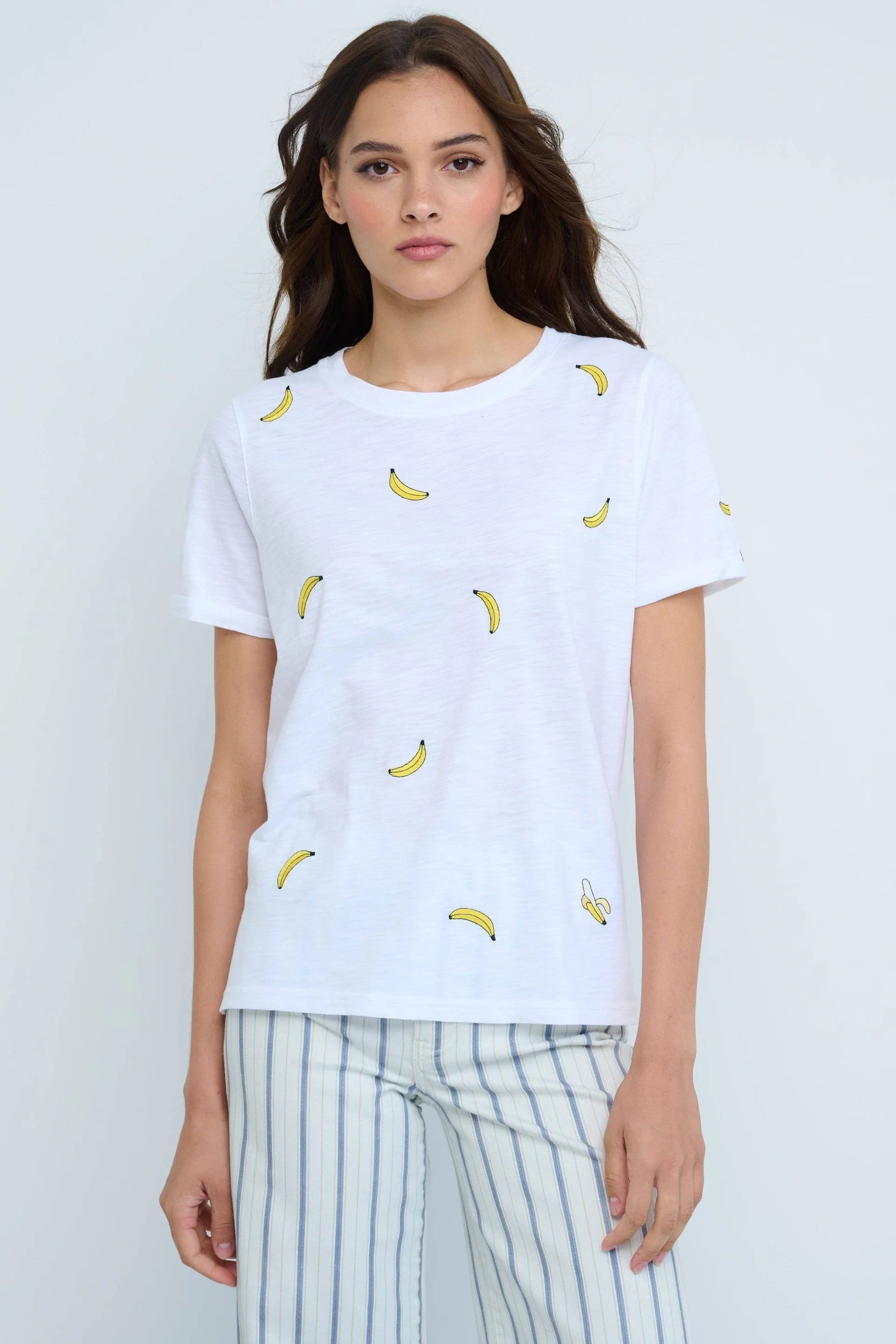 A person wears the Lisa Todd Bananas Tee, featuring small yellow banana embroidery, paired with light striped pants against a plain background for a fresh summer look.