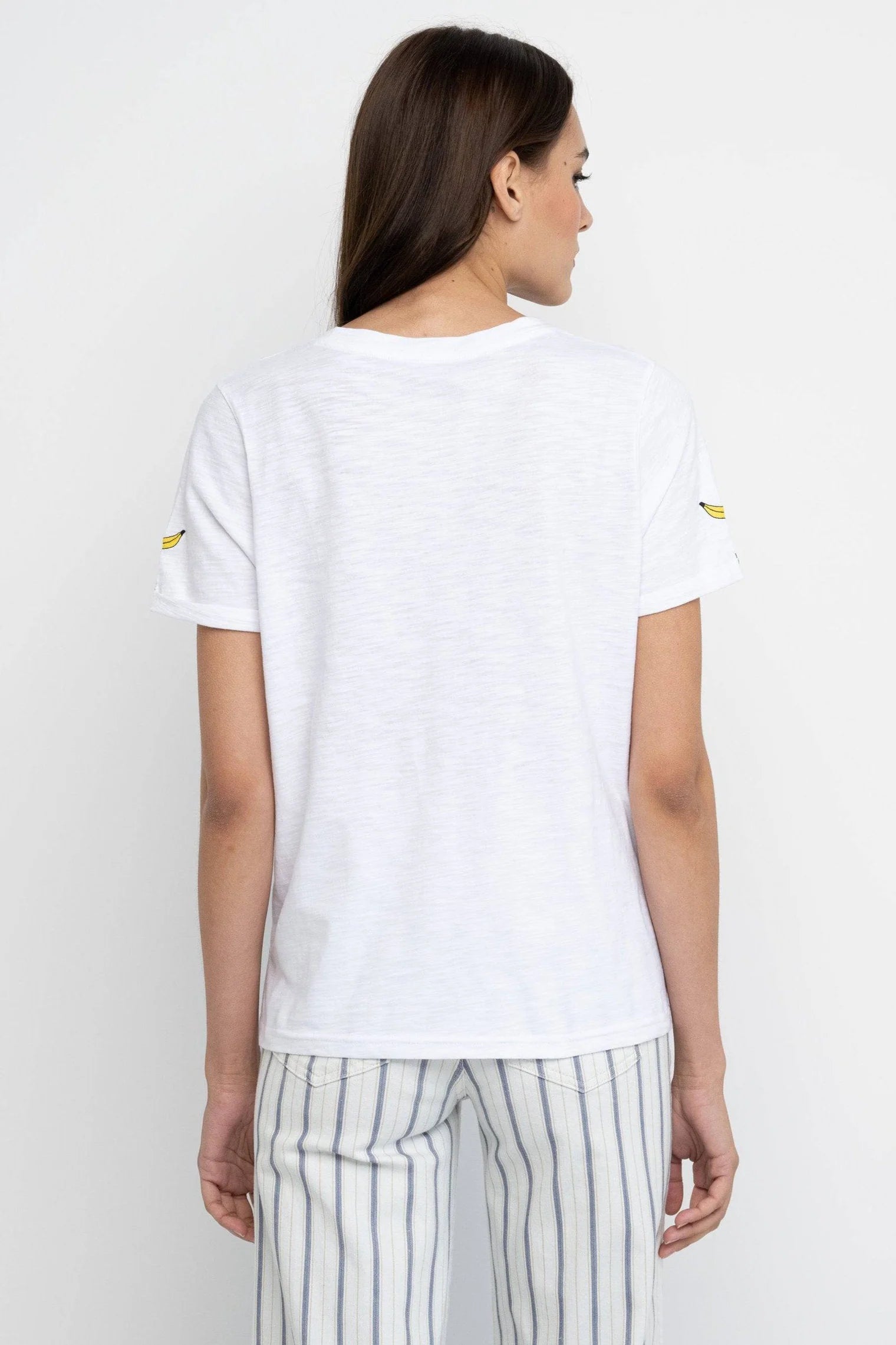 A woman with long brown hair stands with her back to the camera, wearing a Lisa Todd Bananas Tee and white pants with blue and beige stripes, creating a fresh summer look against a light background.