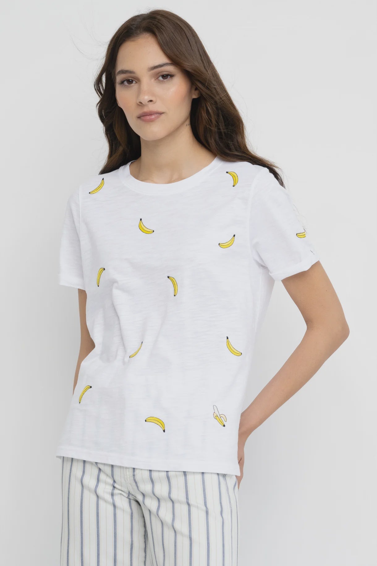 A woman with long brown hair wears the Lisa Todd Bananas Tee, a relaxed fit white T-shirt featuring small yellow banana embroidery, paired with light-colored striped pants. She stands against a plain white background, smiling slightly at the camera.