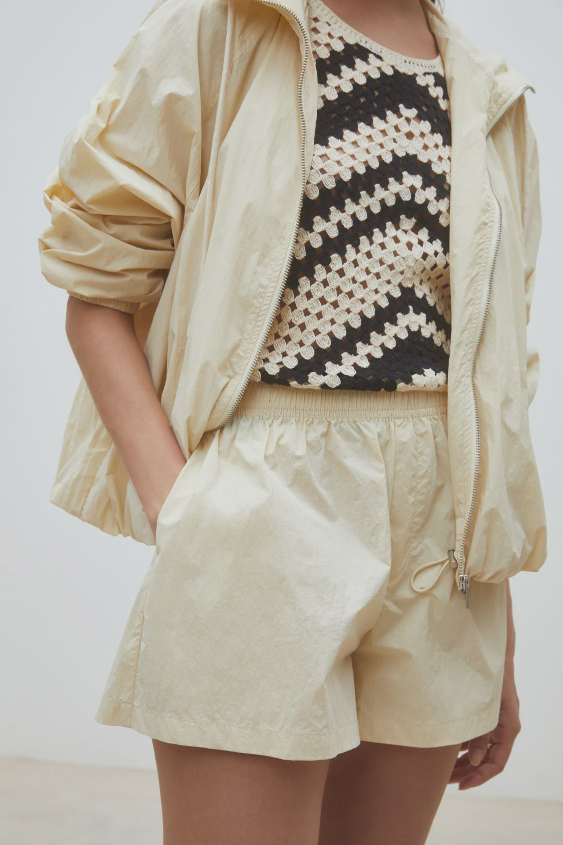 Wearing the SUNCOO Boulbi Short with an elastic waist, a light beige windbreaker, and a black-and-white crocheted zigzag top, this person channels a sporty vibe, left hand casually tucked in their shorts pocket.