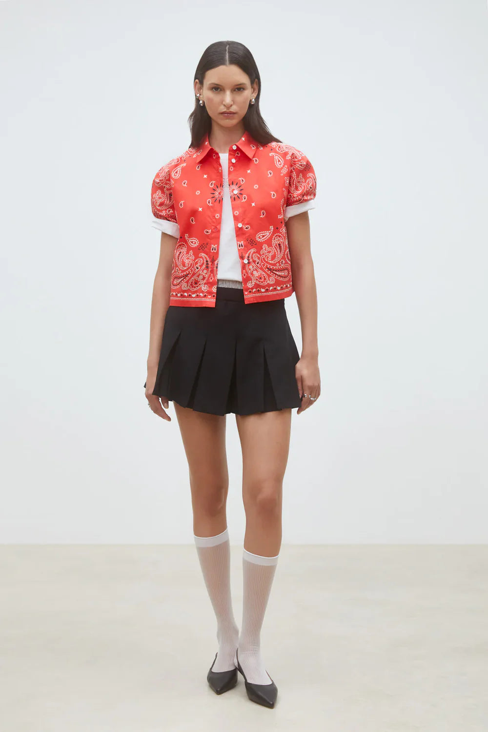 A woman stands against a plain background wearing the SUNCOO Woven Blouse Luz in red, paired with a white top, black pleated mini skirt, long white socks, and black pointed-toe flats.