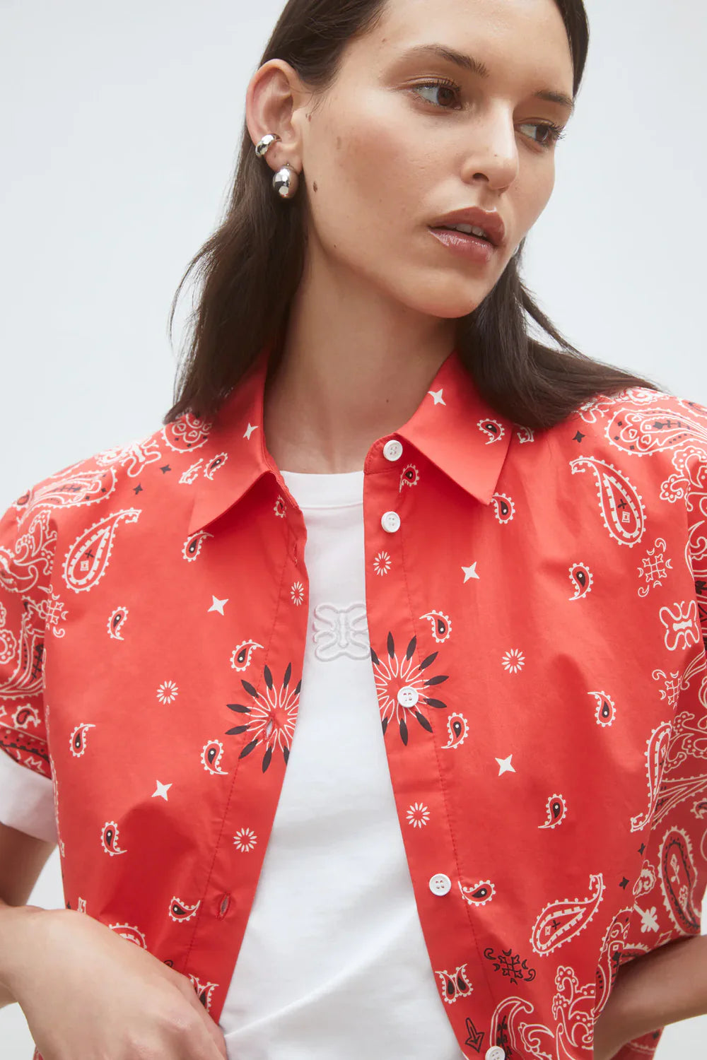 A woman wears the SUNCOO Woven Blouse Luz, featuring a red, white, and black paisley pattern, layered over a white T-shirt. She has long brown hair, pearl earrings, and looks to the side against a plain background.