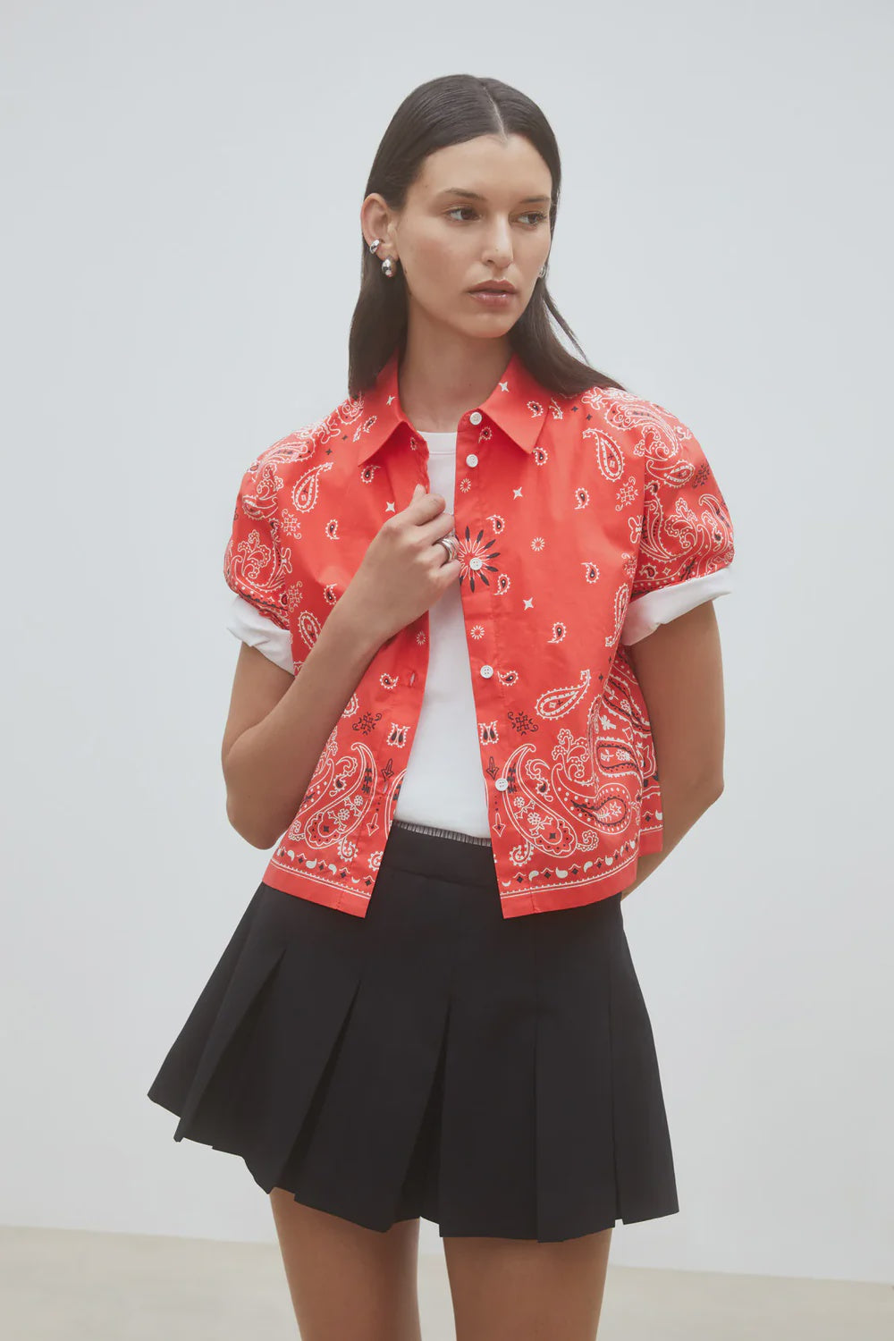 A woman wearing the SUNCOO Woven Blouse Luz stands against a plain light background, layered over a white shirt and paired with a black pleated mini skirt, looking slightly to the side.