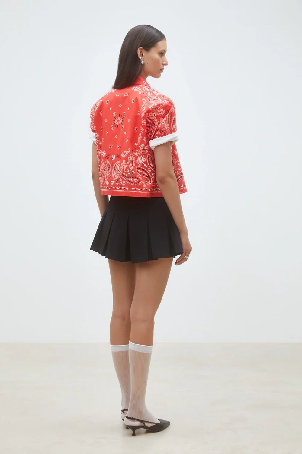 A woman faces sideways in a SUNCOO Woven Blouse Luz with a red bandana pattern, styled with a black pleated mini skirt, sheer white knee-high socks, and black Mary Jane shoes against a plain light background.