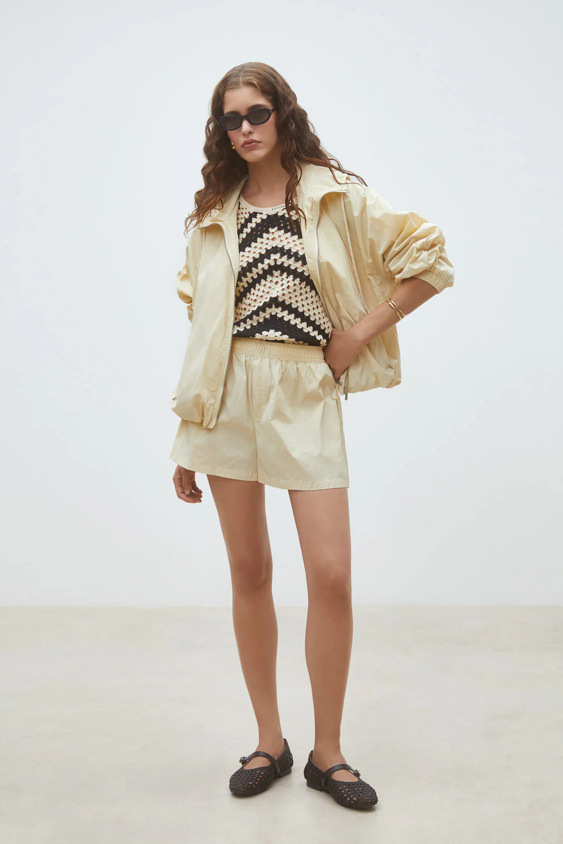 A woman with wavy hair stands against a plain background wearing the SUNCOO Derek Jacket and matching shorts, paired with a black and beige zigzag-patterned top, black sunglasses, and shoes. Her hands are in her pockets.