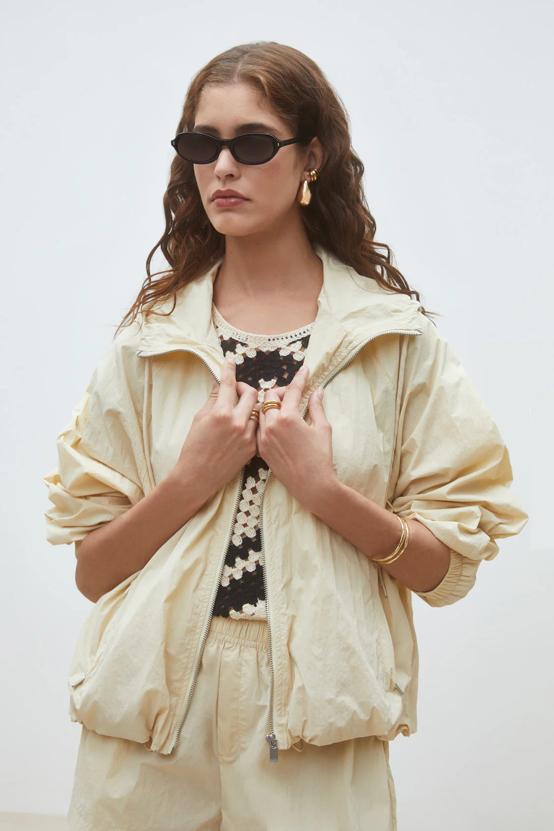 A woman in the SUNCOO Derek Jacket, matching pants, a black and white floral crochet top, black sunglasses, and gold jewelry stands against a light background. She has wavy brown hair and a neutral expression. Stylish weather protection.