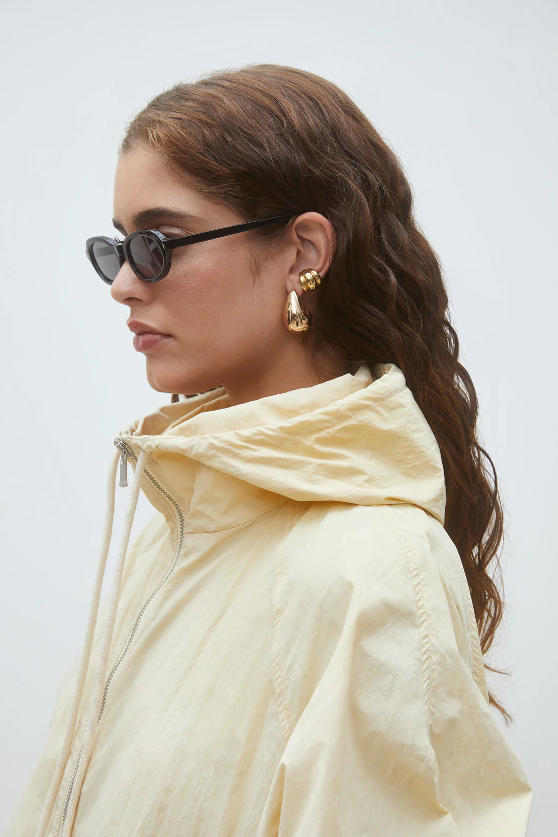 A woman with wavy brown hair wears black oval sunglasses, gold statement earrings, and the SUNCOO Derek Jacket—a light yellow windbreaker with a high collar—against a plain light background.