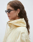 A woman with wavy brown hair wears black oval sunglasses, gold statement earrings, and the SUNCOO Derek Jacket—a light yellow windbreaker with a high collar—against a plain light background.