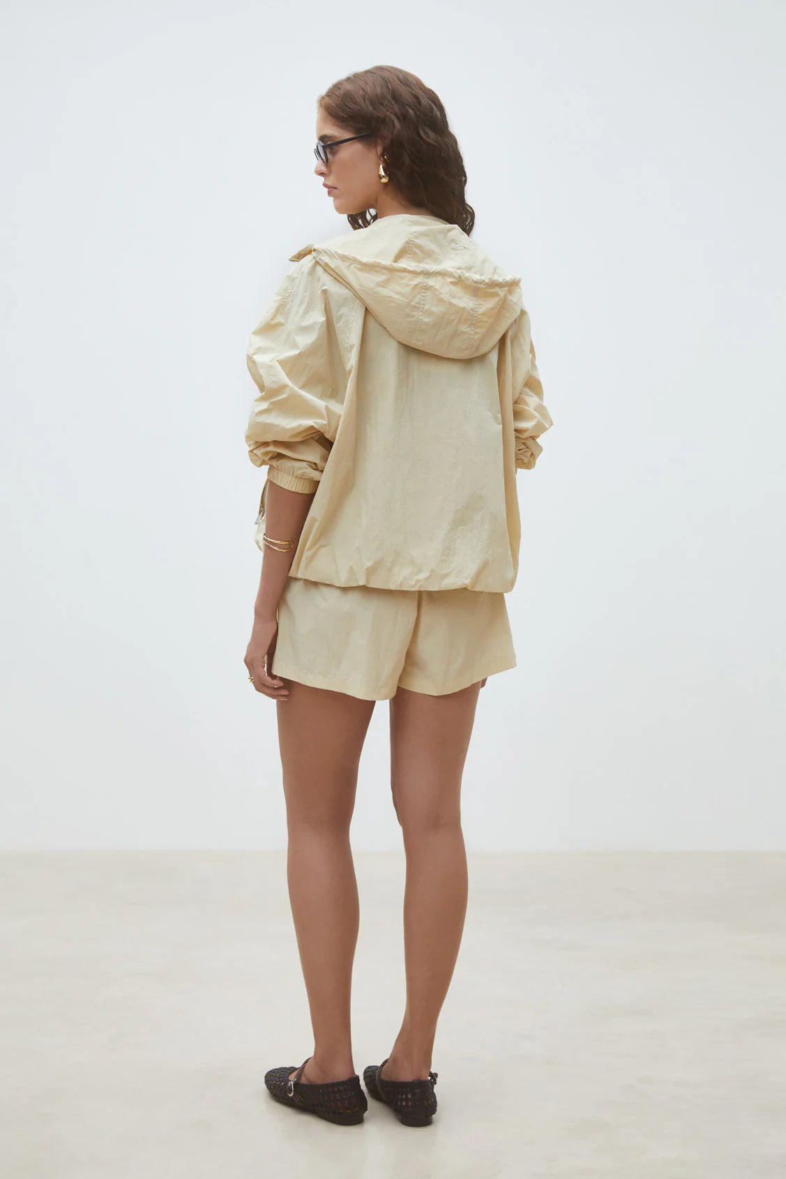 A woman with long wavy hair and glasses stands against a white background, wearing the SUNCOO Derek Jacket in light beige with matching shorts and black slip-on shoes.