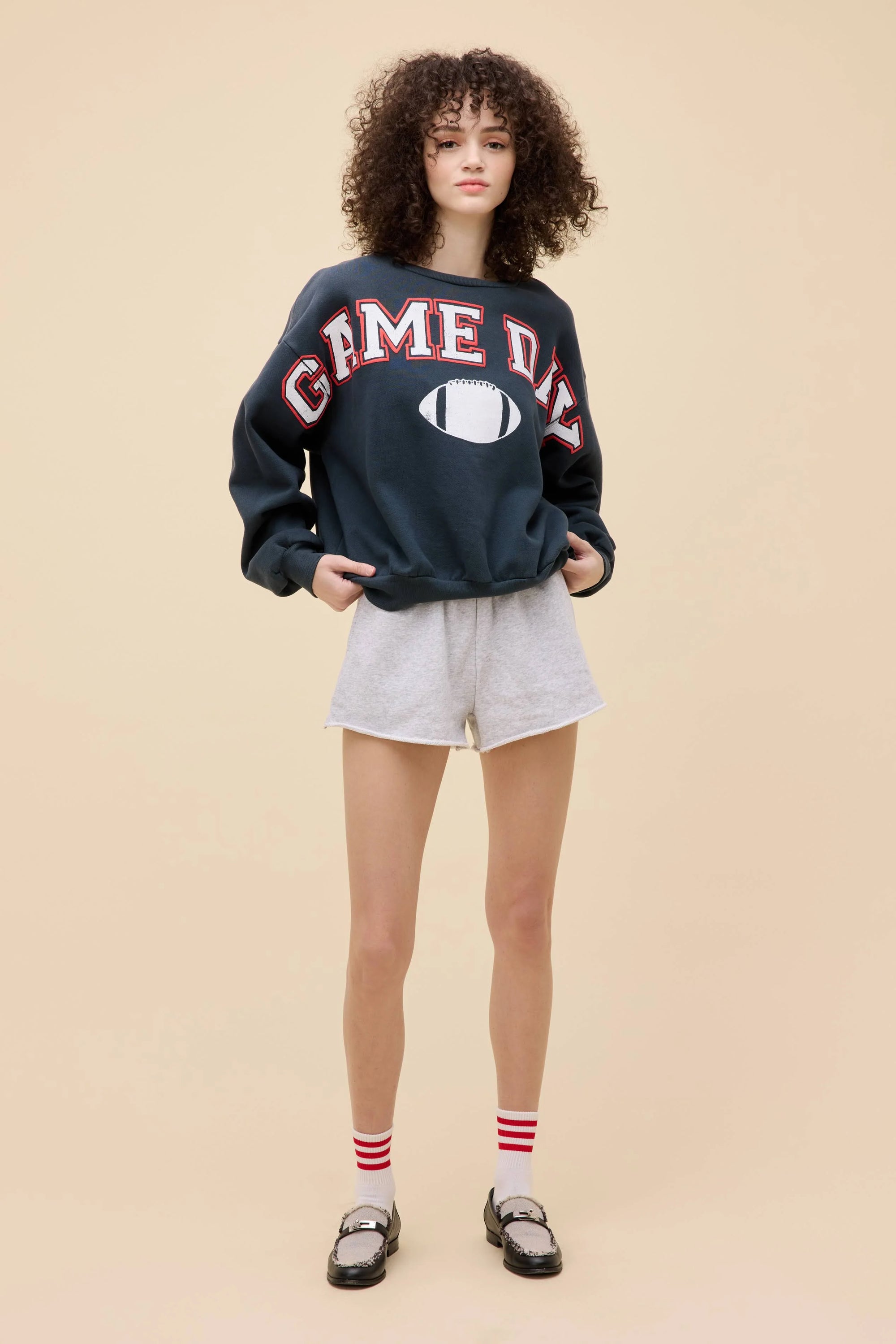 A person with curly hair stands against a beige background, wearing the DayDreamer Game Day Sweatshirt with a football graphic, light gray shorts, red-and-white striped socks, and loafers. Their hands are in their pockets.