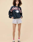 A person with curly hair stands against a beige background, wearing the DayDreamer Game Day Sweatshirt with a football graphic, light gray shorts, red-and-white striped socks, and loafers. Their hands are in their pockets.
