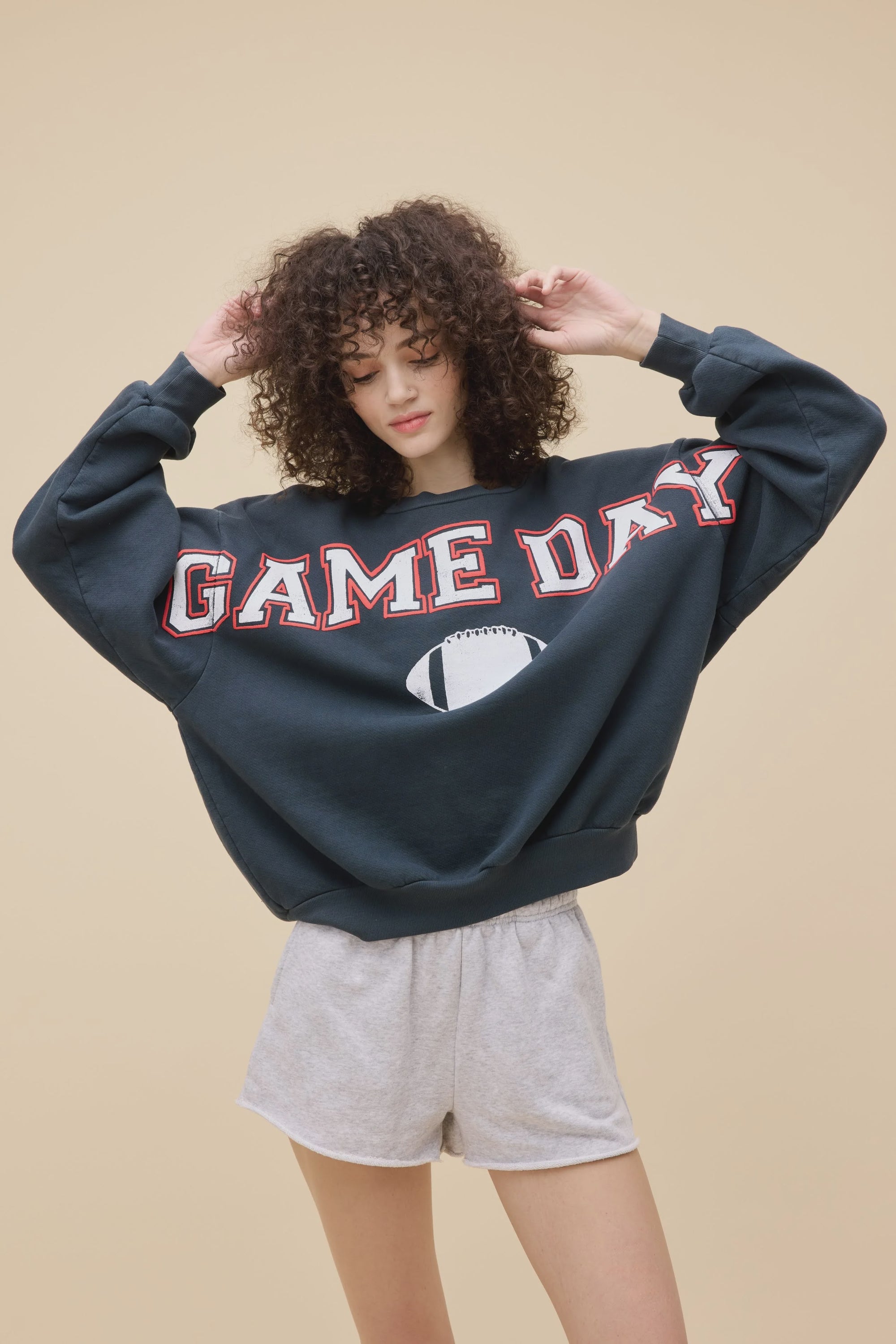 A curly-haired person wears the DayDreamer Game Day Sweatshirt, a dark fleece crew with a football graphic, paired with light gray shorts, standing against a plain beige background.