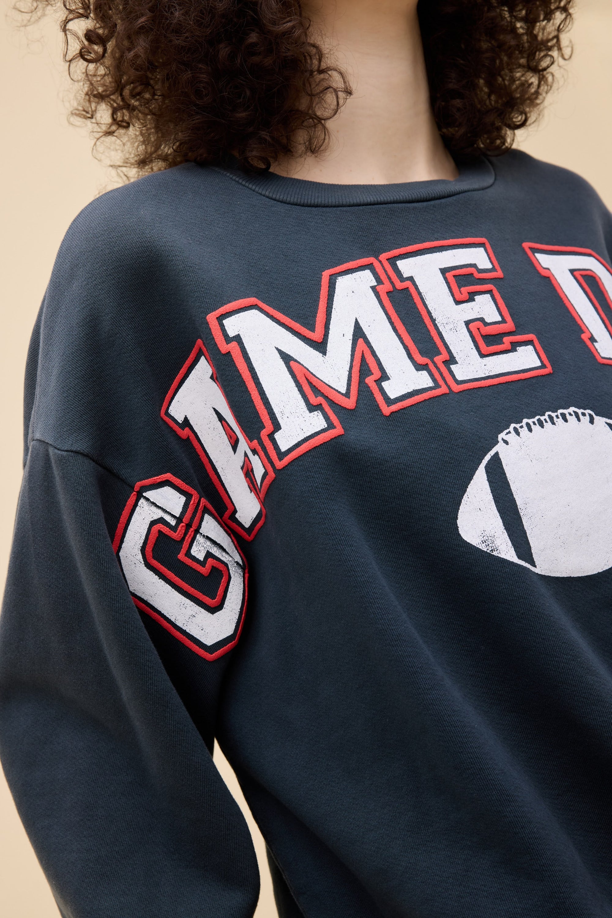 A person with curly hair wears the DayDreamer Game Day Sweatshirt, a cozy fleece crew with bold white and red “GAME D” lettering and a football graphic, set against a plain beige background—ideal for showing off Game Day style.