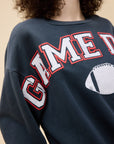 A person with curly hair wears the DayDreamer Game Day Sweatshirt, a cozy fleece crew with bold white and red “GAME D” lettering and a football graphic, set against a plain beige background—ideal for showing off Game Day style.