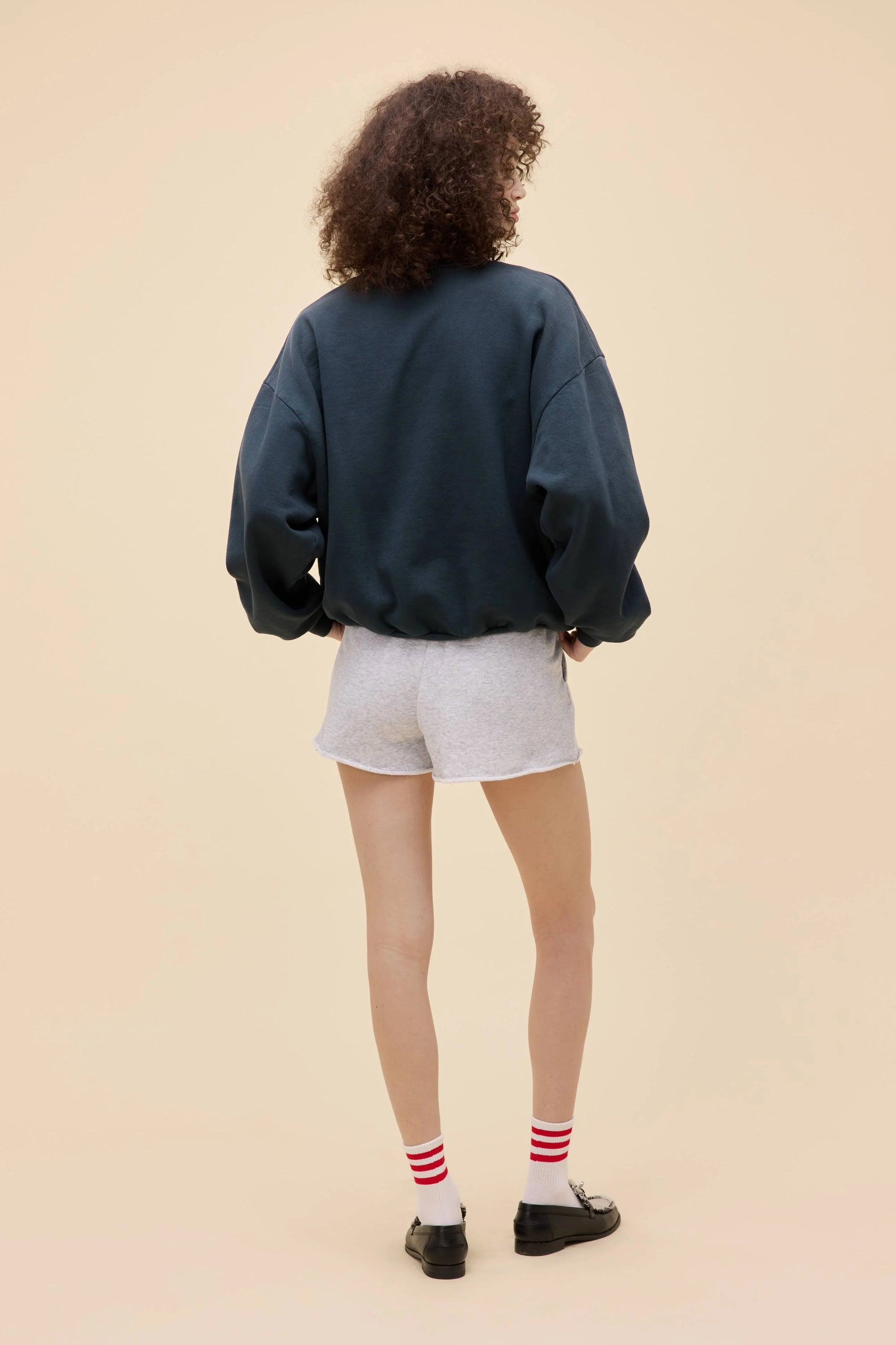 Facing away, a person with curly hair wears the DayDreamer Game Day Sweatshirt in a dark, oversized boxy fit, paired with light gray shorts, red and white striped socks, and black shoes against a plain beige background.