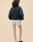 Facing away, a person with curly hair wears the DayDreamer Game Day Sweatshirt in a dark, oversized boxy fit, paired with light gray shorts, red and white striped socks, and black shoes against a plain beige background.
