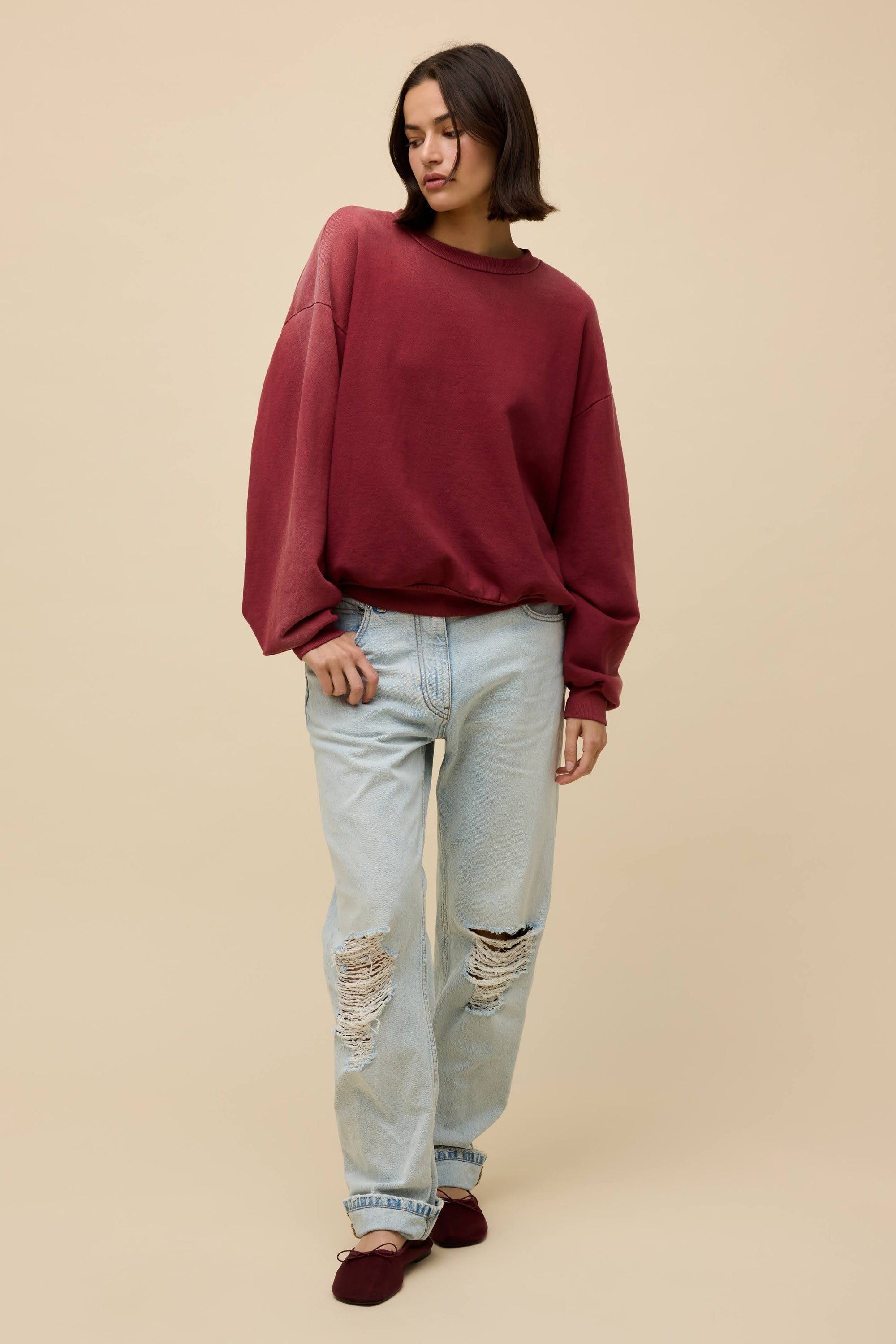 A person with short dark hair wears a maroon DayDreamer Crew Sweatshirt, light blue ripped jeans, and maroon shoes while standing against a plain beige background.