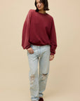 A person with short dark hair wears a maroon DayDreamer Crew Sweatshirt, light blue ripped jeans, and maroon shoes while standing against a plain beige background.