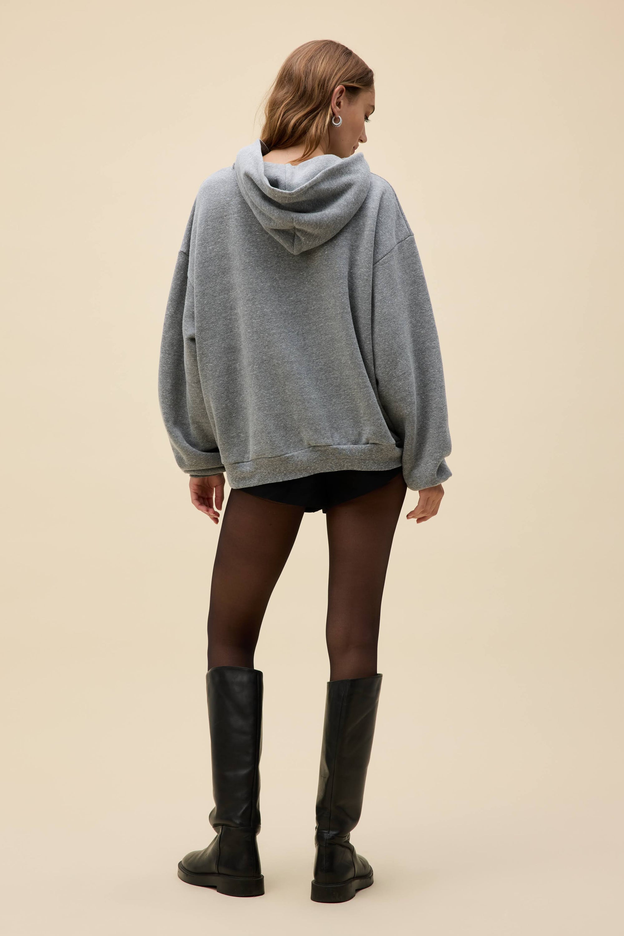 A person stands with their back to the camera in a loose DayDreamer FleetWood Mac Hoodie, black shorts, sheer black tights, and knee-high black boots against a plain beige background.