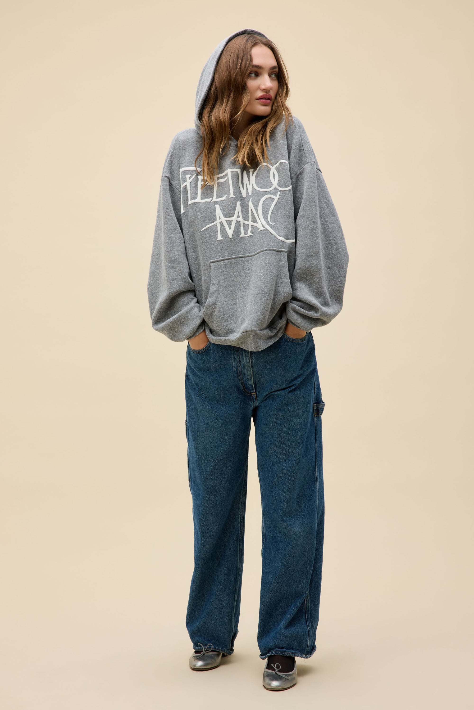A woman wears a DayDreamer FleetWood Mac Hoodie with the hood up, paired with loose blue jeans and black sneakers, standing against a plain beige background and looking to the side with her hands in her pockets.