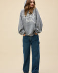 A woman wears a DayDreamer FleetWood Mac Hoodie with the hood up, paired with loose blue jeans and black sneakers, standing against a plain beige background and looking to the side with her hands in her pockets.