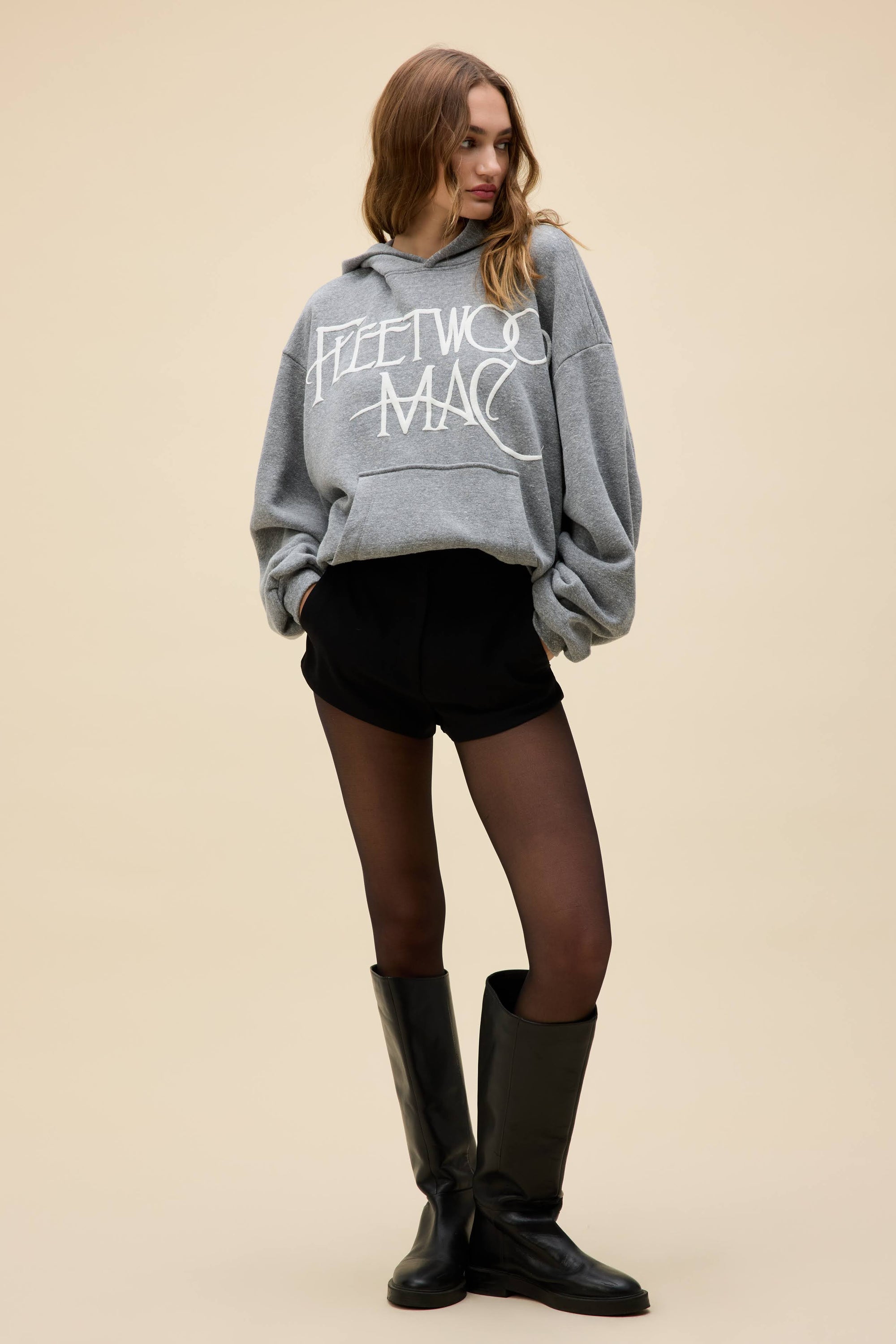 A person wears an oversized DayDreamer FleetWood Mac Hoodie, black shorts, sheer black tights, and black knee-high boots while standing against a plain beige background.