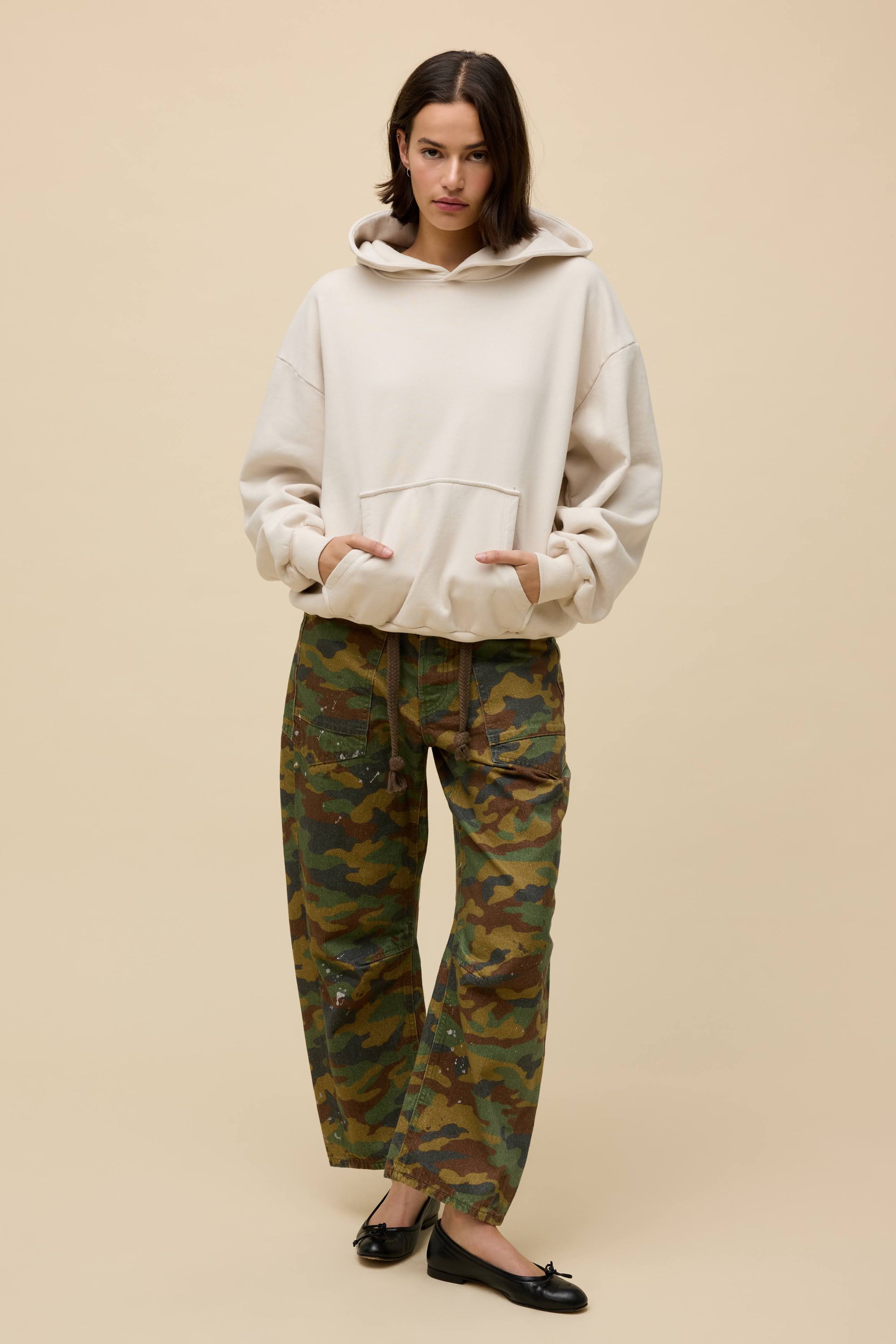 A woman stands against a beige background wearing the DayDreamer OS Hoodie, paired with camouflage pants and black ballet flats, her hands in the hoodie pocket and expression neutral.